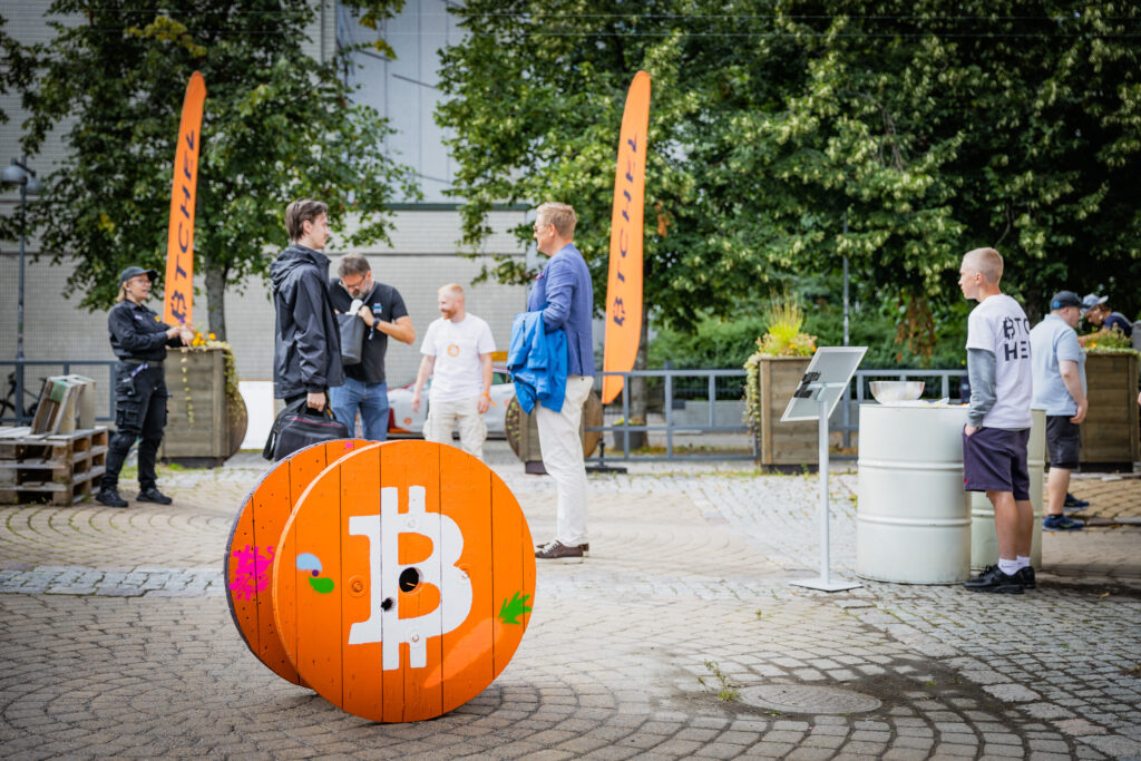 Entrance area of the BTCHEL Bitcoin conference in Helsinki, with attendees chatting near bright orange Bitcoin-themed decor and BTCHEL flags, capturing the event’s vibrant atmosphere.